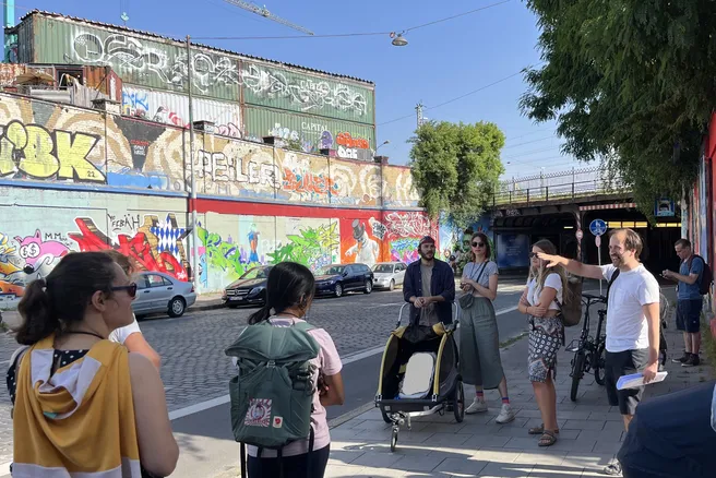 People are standing on the pavement and there is a lot of graffiti behind them.