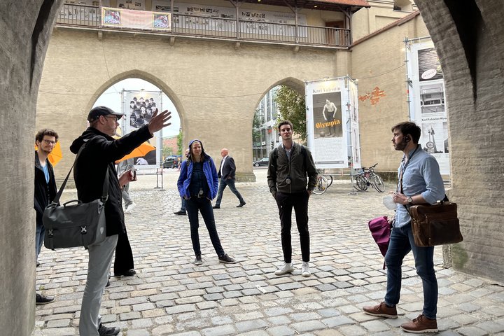 People are standing in a courtyard listening to a person pointing upwards with his hand. 