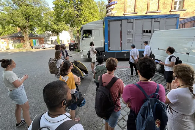 People are standing next to a road on which a cattle truck is passing.