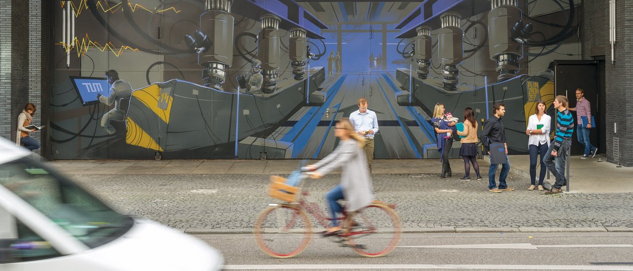 Vordergrund Auto und Fahrradfahrerin; Hintergrund Studierende vor dem Campus Innenstadt