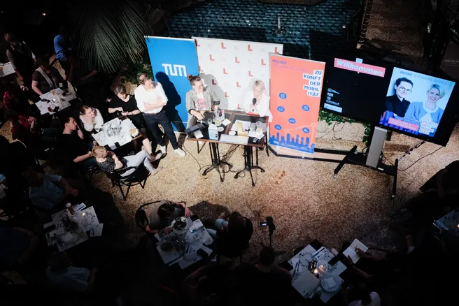 3 People sit on higher chairs in front on a audience. 